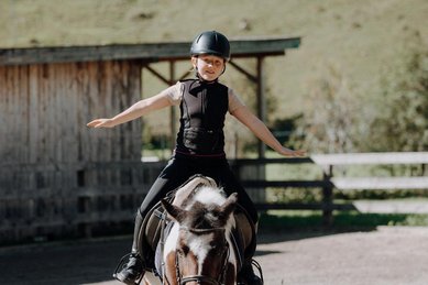 Ein Kind sitzt mit ausgebreiteten Armen auf einem Pony im Außenbereich des Familienhotels Oberkarteis im Salzburger Land.