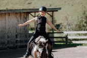 Ein Kind sitzt mit ausgebreiteten Armen auf einem Pony im Außenbereich des Familienhotels Oberkarteis im Salzburger Land.