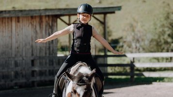 Ein Kind sitzt mit ausgebreiteten Armen auf einem Pony im Außenbereich des Familienhotels Oberkarteis im Salzburger Land.