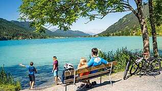 Familie macht eine Pause vom Fahrradfahren am See. Die Eltern sitzen auf einer Bank und die Kinder spielen am Wasser.