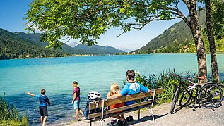 Familie macht eine Pause vom Fahrradfahren am See. Die Eltern sitzen auf einer Bank und die Kinder spielen am Wasser.