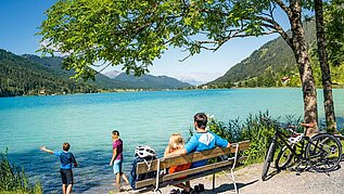 Familie macht eine Pause vom Fahrradfahren am See. Die Eltern sitzen auf einer Bank und die Kinder spielen am Wasser.