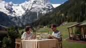 Eltern sitzen bei einer Mahlzeit im Außenbereich des Restaurants und genießen den Blick auf die Berge im Hotel Bella Vista in Südtirol.