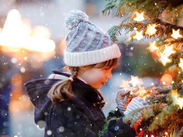 Kind schmückt einen beleuchteten Weihnachtsbaum im Schnee, warm eingepackt in Winterjacke und Strickmütze.
