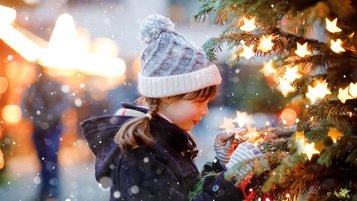 Kind schmückt einen beleuchteten Weihnachtsbaum im Schnee, warm eingepackt in Winterjacke und Strickmütze.