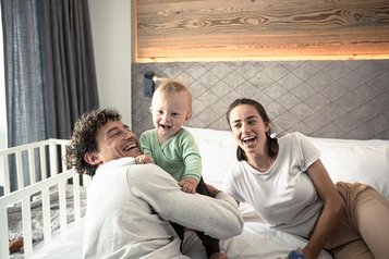 Ein Mann und eine Frau liegen auf dem Bett im Familienhotel und hält sein lachendes Baby im Arm.
