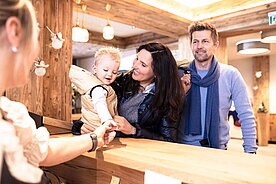 Baby sitzt lachend mit seinen Eltern an der Rezeption eines Familienhotels 