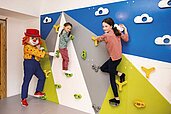 Kinder klettern an der Kletterwand im Happy-Club des Familienhotels Alpenhotel Kindl in Tirol. Auch Clown Happy, das Maskottchen des Familienhotels, klettert mit und bringt die Kinder zum Lachen.