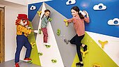 Kinder klettern an der Kletterwand im Happy-Club des Familienhotels Alpenhotel Kindl in Tirol. Auch Clown Happy, das Maskottchen des Familienhotels, klettert mit und bringt die Kinder zum Lachen.