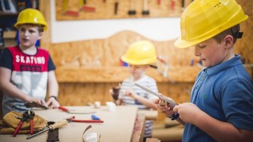 Mehrere Kinder tragen gelbe Schutzhelme und arbeiten in der Werkstatt des Familienhotels Landgut Furtherwirt in Tirol mit Werkzeug und Holz.
