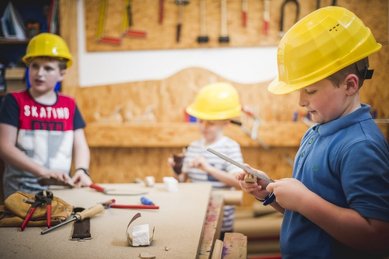 Mehrere Kinder tragen gelbe Schutzhelme und arbeiten in der Werkstatt des Familienhotels Landgut Furtherwirt in Tirol mit Werkzeug und Holz.