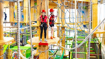 Zwei Kinder in Sicherheitsausrüstung gehen über Holzplattformen in einem bunten Indoor-Hochseilgarten im Hotel Feldberger Hof, umgeben von Seilen und Netzen.
