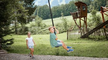 Junge saust mit dem Flying Fox durch den Waldspielplatz des Familienhotels Krone im Allgäu, während ein Mädchen zuschaut.