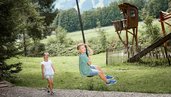 Junge saust mit dem Flying Fox durch den Waldspielplatz des Familienhotels Krone im Allgäu, während ein Mädchen zuschaut.
