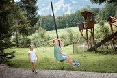 Junge saust mit dem Flying Fox durch den Waldspielplatz des Familienhotels Krone im Allgäu, während ein Mädchen zuschaut.