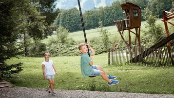 Junge saust mit dem Flying Fox durch den Waldspielplatz des Familienhotels Krone im Allgäu, während ein Mädchen zuschaut.
