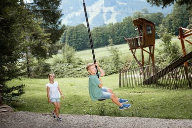Junge saust mit dem Flying Fox durch den Waldspielplatz des Familienhotels Krone im Allgäu, während ein Mädchen zuschaut.