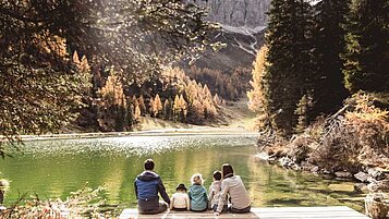 Eine Familie mit drei Kindern sitzt im Herbst auf einem Holzsteg und blickt auf den See und die Berge.