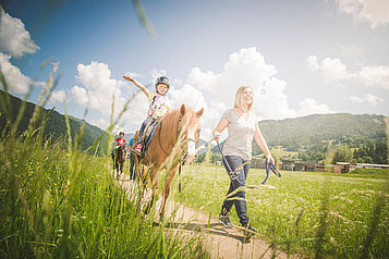 Reitlehrerin führt ein Mädchen auf einem Pferd durch die Wanderwege nahe des Familienhotels Landgut Furtherwirt in Tirol.