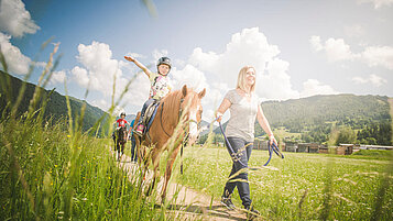 Reitlehrerin führt ein Mädchen auf einem Pferd durch die Wanderwege nahe des Familienhotels Landgut Furtherwirt in Tirol.