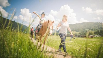 Reitlehrerin führt ein Mädchen auf einem Pferd durch die Wanderwege nahe des Familienhotels Landgut Furtherwirt in Tirol.