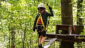 Junge spaziert in Kletterausrüstung über einen Holzpfad im Klettergarten.