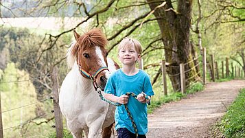 Ein Kind führt ein Pony an einer Leine durch einen Wald.