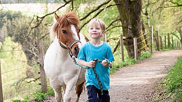 Ein Kind führt ein Pony an einer Leine durch einen Wald.