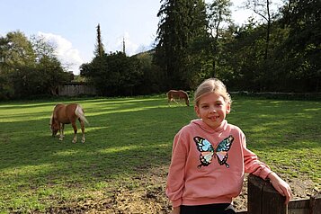 Ein Mädchen sitzt auf dem Zaun und im Hintergrund laufen die Pferde auf der Wiese im Familienhotel Monikas Ferienhof.