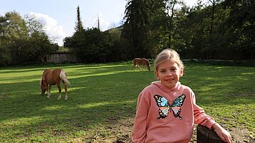 Ein Mädchen sitzt auf dem Zaun und im Hintergrund laufen die Pferde auf der Wiese im Familienhotel Monikas Ferienhof.