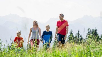 Eine Familie mit zwei Kindern genießt eine Wanderung in der idyllischen Allgäuer Berglandschaft, umgeben von grünen Wiesen und Berggipfeln im Hintergrund, symbolisch für Urlaub im Familienhotel Allgäuer Berghof.