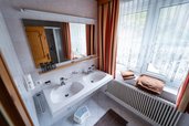 Helles Badezimmer im Familienhotel Bruckwirt mit Doppelwaschbecken, Spiegel und großen Fenstern.