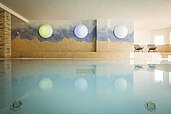 Entspannendes Hallenbad im Familienhotel mit ruhigem Wasser, gestaltet mit einer künstlerischen Wand mit Bergen und schwebenden Kugellampen, begleitet von stilvollen Liegestühlen am Rand des Beckens.