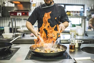 Koch in Aktion beim Flambieren eines Gerichts in einer professionellen Küche, mit hohen Flammen über einer Pfanne, was auf eine dynamische und geschickte Zubereitung hinweist.