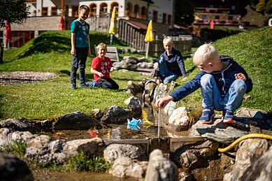 Kinder spielen auf dem Spielplatz am kleinen Wasserbach und lassen Schiffchen treiben im Familienhotel Oberkarteis im Salzburger Land.