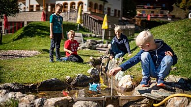 Kinder spielen auf dem Spielplatz am kleinen Wasserbach und lassen Schiffchen treiben im Familienhotel Oberkarteis im Salzburger Land.