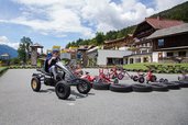 Ein Kind fährt mit einem Gokart auf einer Outdoor-Bahn, umgeben von Spielgeräten und Tricycles, mit dem Kärntner Familienhotel Ramsi im Hintergrund