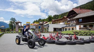 Ein Kind fährt mit einem Gokart auf einer Outdoor-Bahn, umgeben von Spielgeräten und Tricycles, mit dem Kärntner Familienhotel Ramsi im Hintergrund