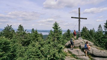 Drei Wanderer an einem Gipfelkreuz auf einer Felskuppe über dichtem Wald mit weiter Aussicht.