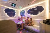Familie sitzt lachend im farbenfroh beleuchteten Dampfbad des Zauchenseehofs, umgeben von spielerischen Wolken- und Tierprojektionen an den Wänden.