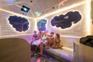 Familie sitzt lachend im farbenfroh beleuchteten Dampfbad des Zauchenseehofs, umgeben von spielerischen Wolken- und Tierprojektionen an den Wänden.