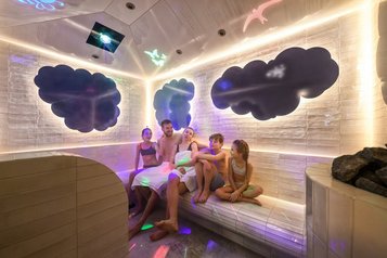 Familie sitzt lachend im farbenfroh beleuchteten Dampfbad des Zauchenseehofs, umgeben von spielerischen Wolken- und Tierprojektionen an den Wänden.