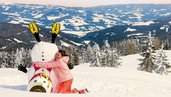 Ein Kind umarmt einen umgefallenen Schneeman in Kaernten. Der Ausblick im Winter kann sich sehen lassen und zeigt ein tolles Panorama.