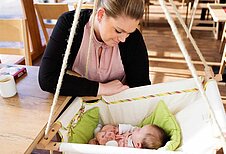 Baby liegt in einer Babyschaukel im Restaurant des Familienhotels Landhuus Laurenz und die Mutter sitzt daneben am Tisch und umsorgt ihr Kind.