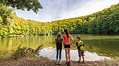Drei Kinder stehen vor einem See und Blicken auf den See und den umliegenden Wald im Sommer.