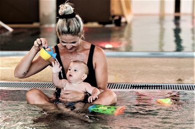 Ein Baby sitzt mit seiner Mutter im Pool des Familienhotel Mia Alpina in Tirol und spielt mit buntem Wasserspielzeug.