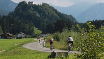 Eine Familie beim Fahrrad fahren im Allgäu. Die Strecke führt auf einem Weg an einem Berg entlang.