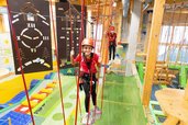 Ein lächelndes Kind in einem roten Helm und Sicherheitsgurt genießt das Klettern in einem Indoor-Spielplatz mit Seilen und Kletterwänden im Hotel Feldberger Hof.