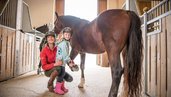 Eine Reitlehrerin und ein Mädchen mit Reithelm stehen neben einem braunen Pferd im Landgut Furtherwirt, während das Kind stolz ein Hufeisen hält