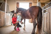 Eine Reitlehrerin und ein Mädchen mit Reithelm stehen neben einem braunen Pferd im Landgut Furtherwirt, während das Kind stolz ein Hufeisen hält